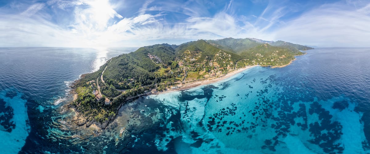 An aerial view of a coastal island with clear blue waters, lush green hills, and a winding road along the coastline.