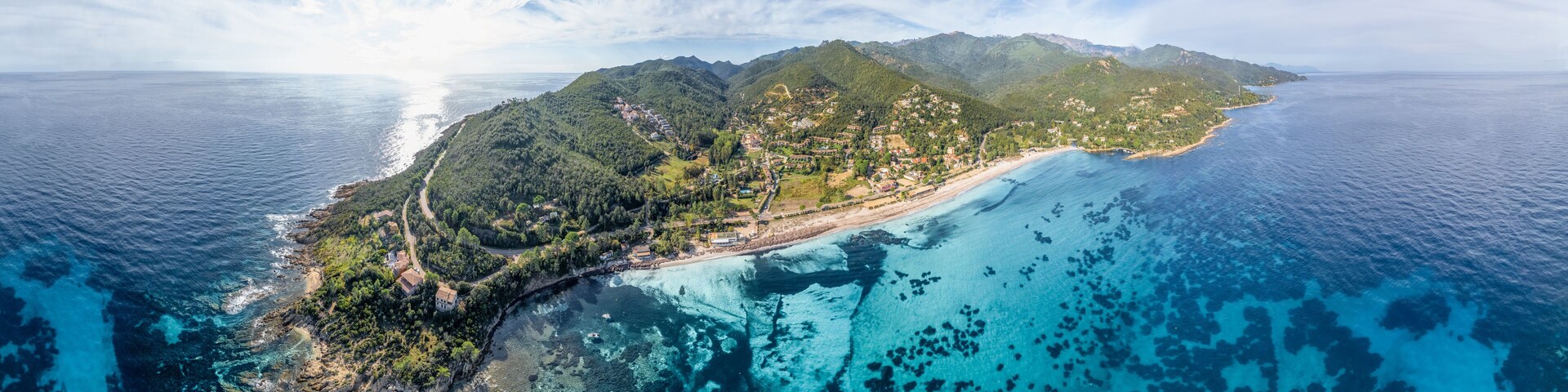 An aerial view of a coastal island with clear blue waters, lush green hills, and a winding road along the coastline.
