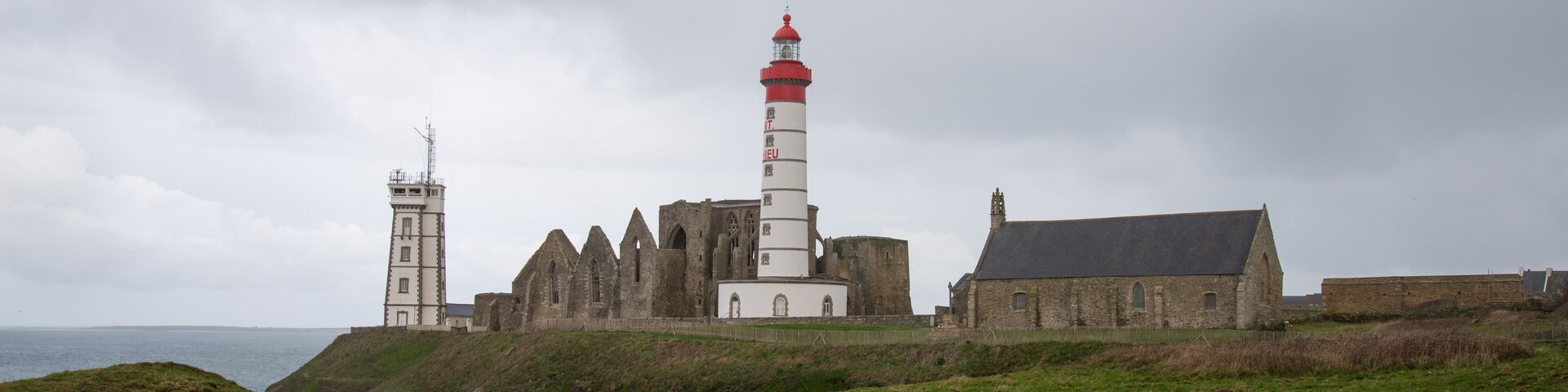 A coastal lighthouse with a red top, a white tower, and a church with a steep roof.