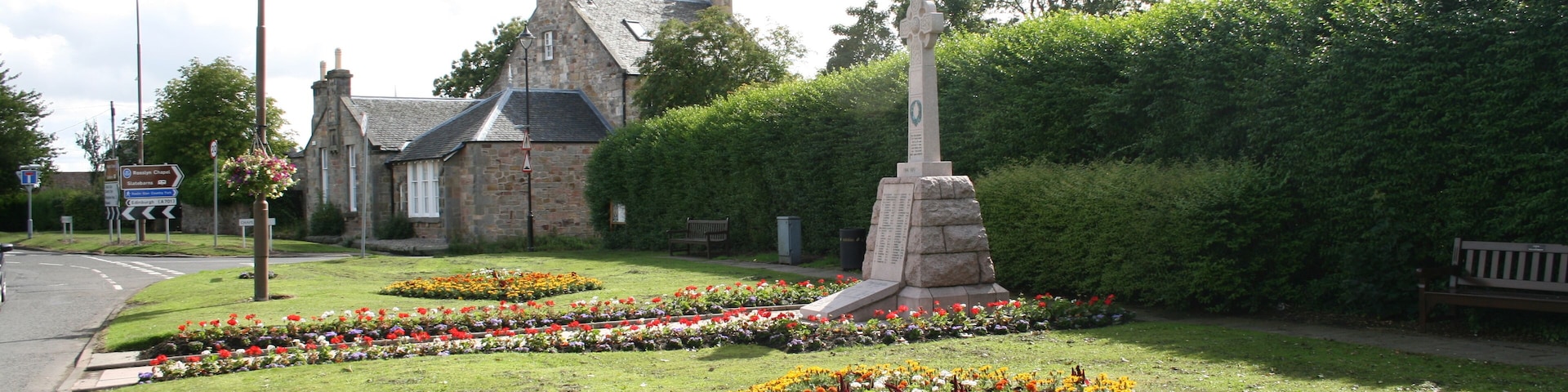 Un césped con un monumento de piedra, macizos de flores, un edificio de piedra y una banca.