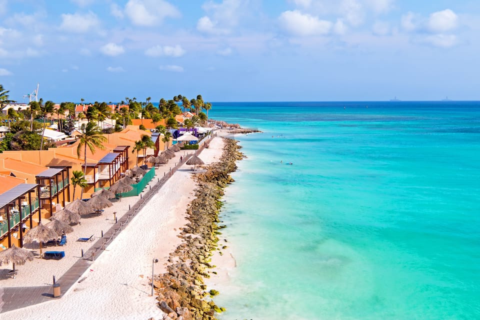 A coastal resort with a white sandy beach, turquoise waters, palm trees, and overwater bungalows.