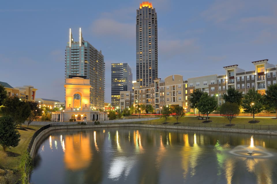 A cityscape with a large body of water reflecting lights, surrounded by modern buildings and a park with trees.