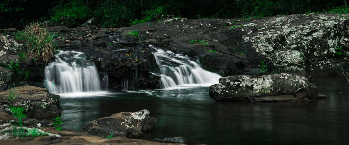 Uma cascata despencando sobre rochas em uma piscina calma, cercada por vegetação.