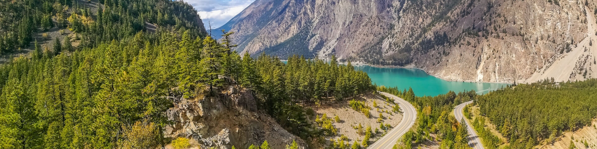 Uma estrada sinuosa serpenteando pelas montanhas, um lago azul-turquesa e montanhas cobertas de floresta.