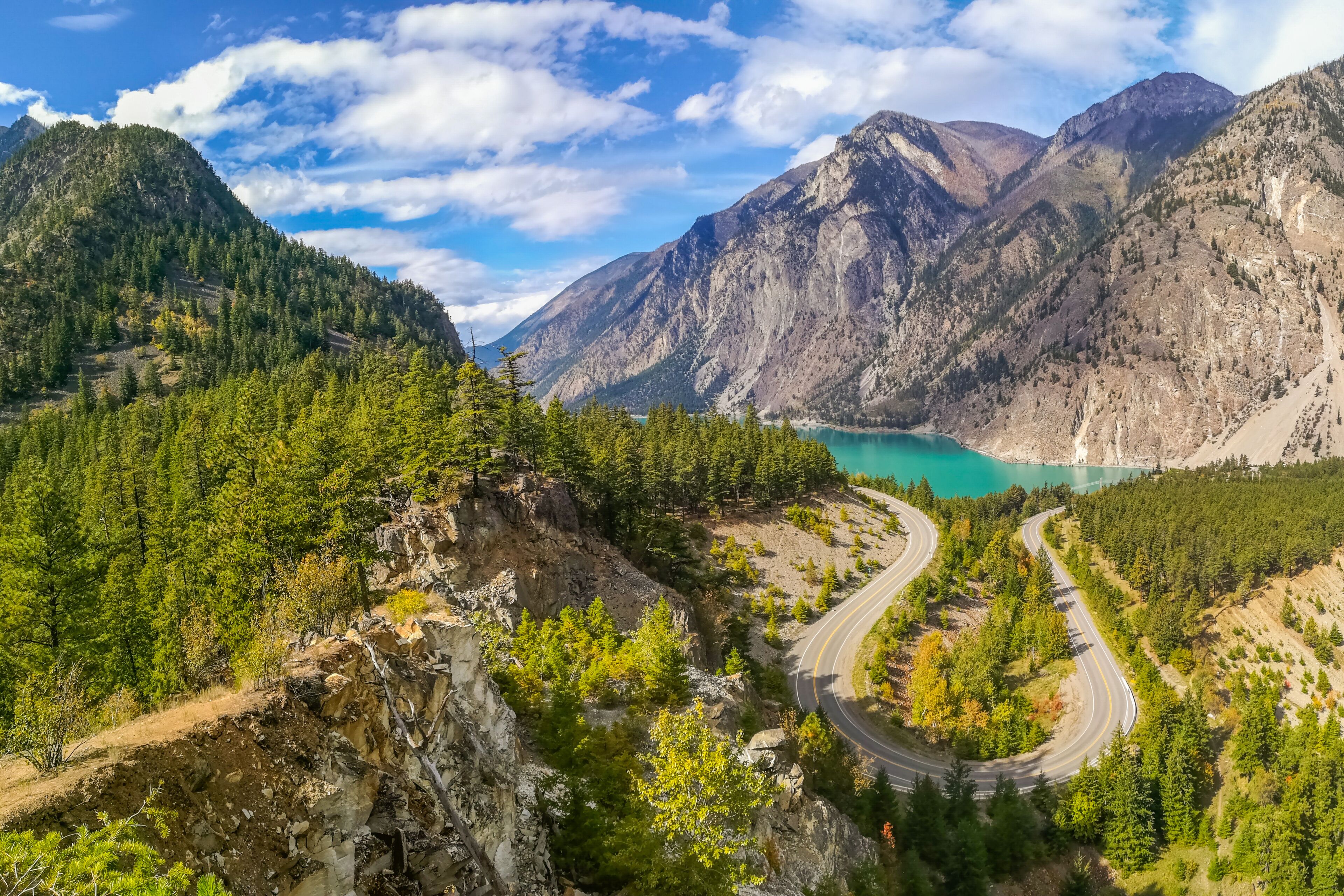A winding mountain road, a turquoise lake, and forested mountains.