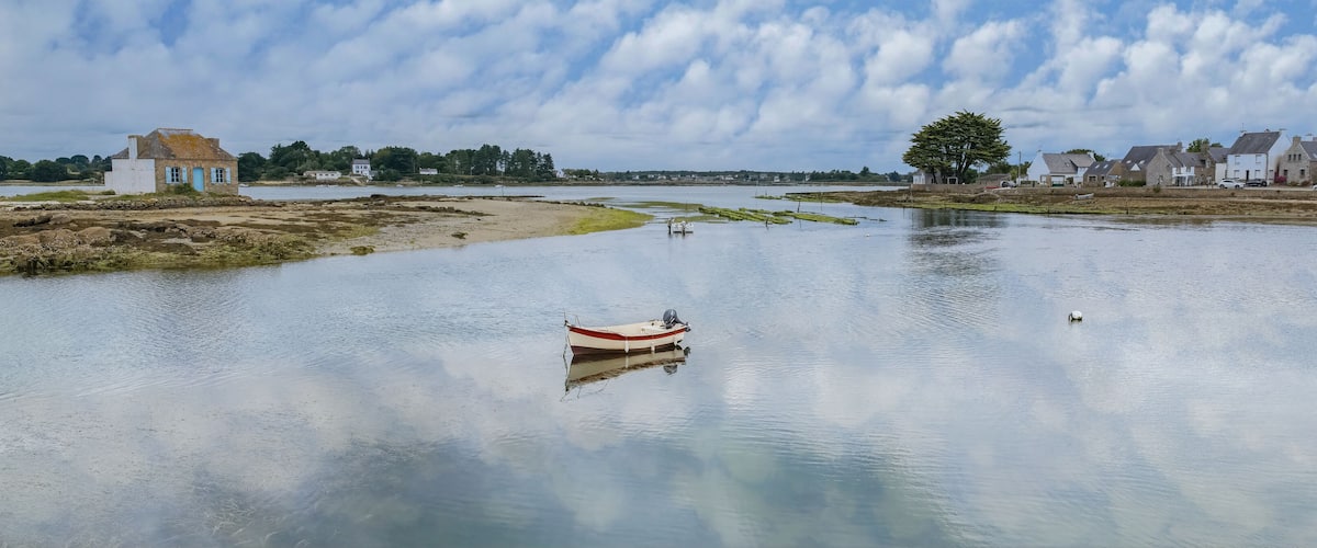 A calm river with a small boat anchored in the middle, surrounded by houses and trees.