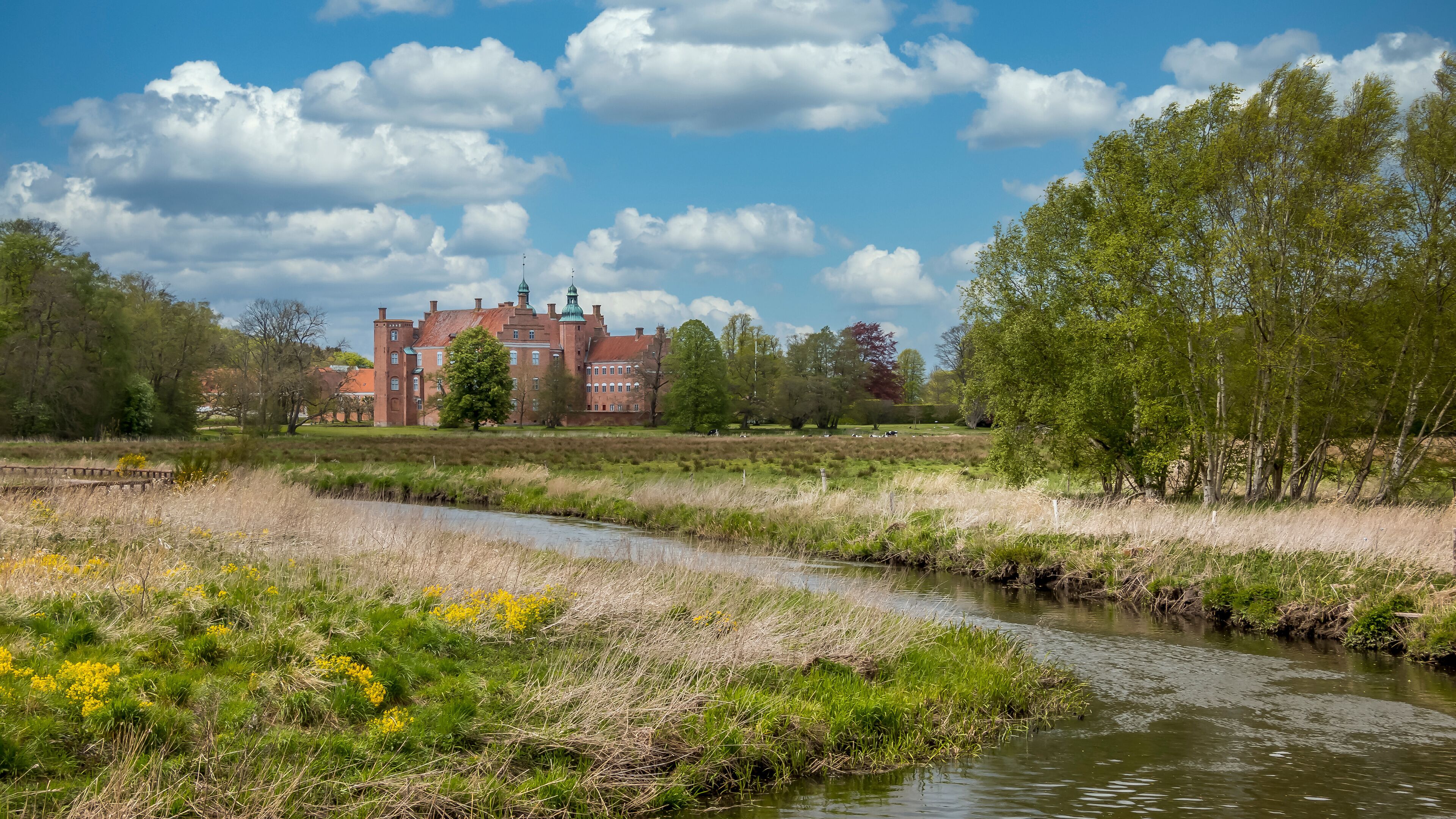 A river meandering through a grassy field with yellow flowers, a brick building with green roofs, and trees in the background.