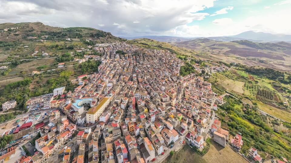 A densely packed town with red-roofed buildings, surrounded by green hills and mountains.