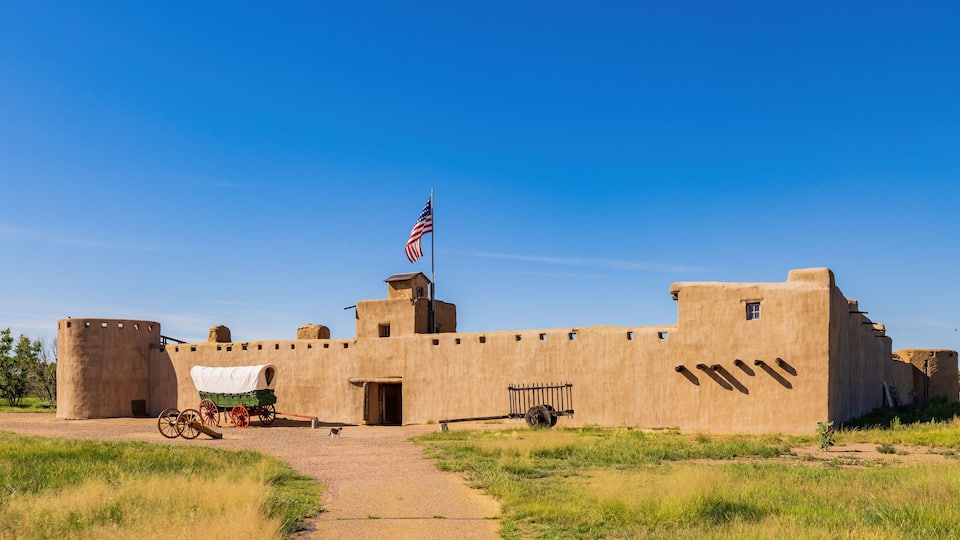 A historic fort with a flag, a covered wagon, and a cart.