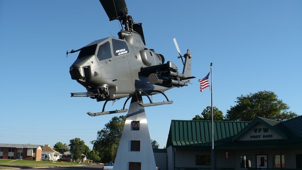 A military helicopter with a flag on top, a monument, and a post office in the background.