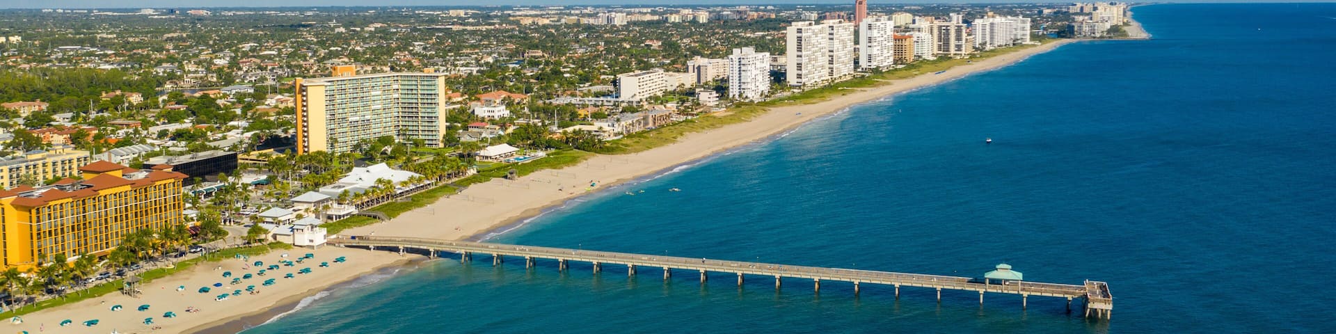 Un paisaje urbano costero con una playa extensa, un muelle que se adentra en el océano y altos edificios a lo largo de la costa.