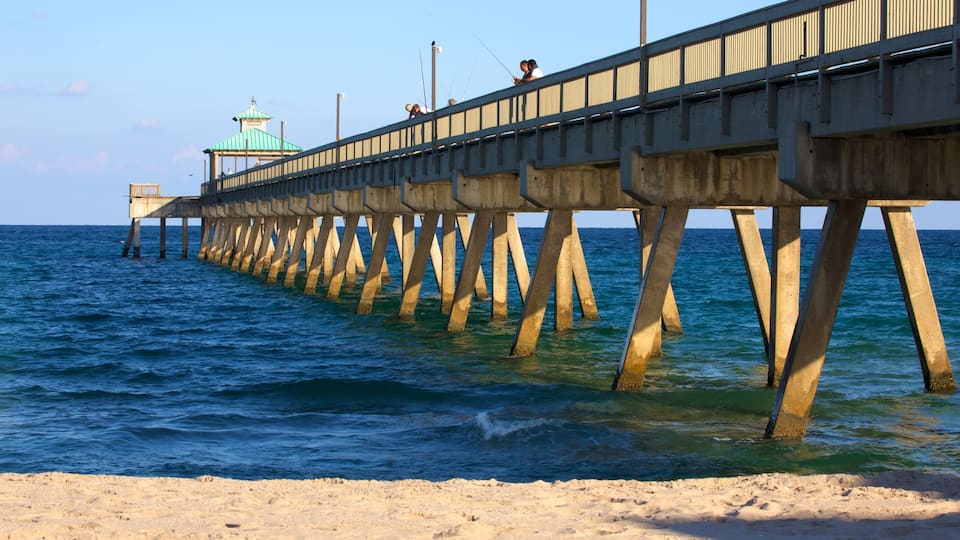 A pier extending into the ocean with people fishing, a lighthouse in the background, and a sandy beach in the foreground.