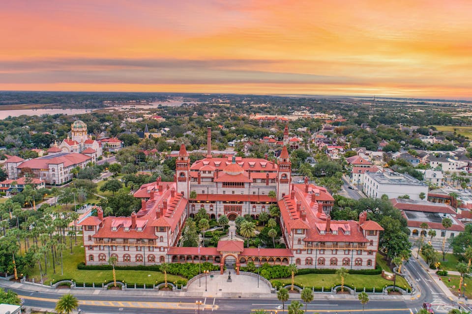 A large, historic building complex with red-tiled roofs, surrounded by lush greenery and a river in the background.