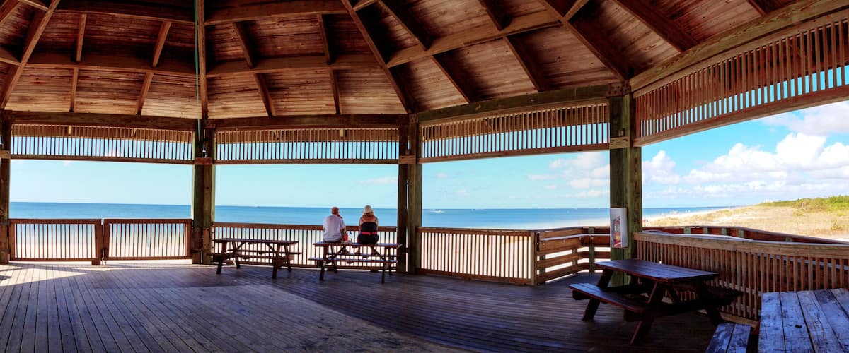 A wooden deck with picnic tables and benches overlooking a beach.