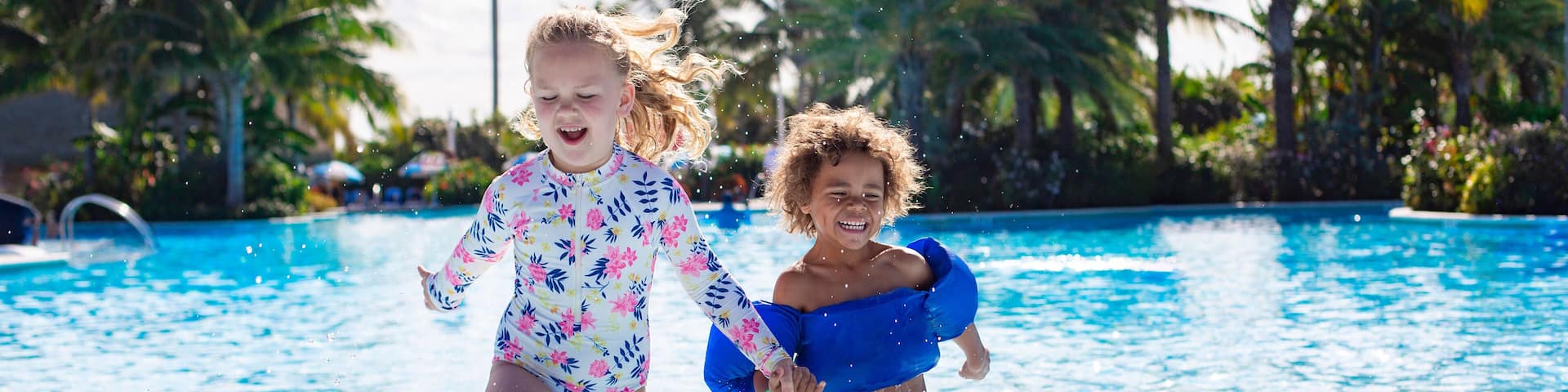 A Cute diverse boy and little girl running and splashing together holding hands in the large swimming pool resort while on a family vacation