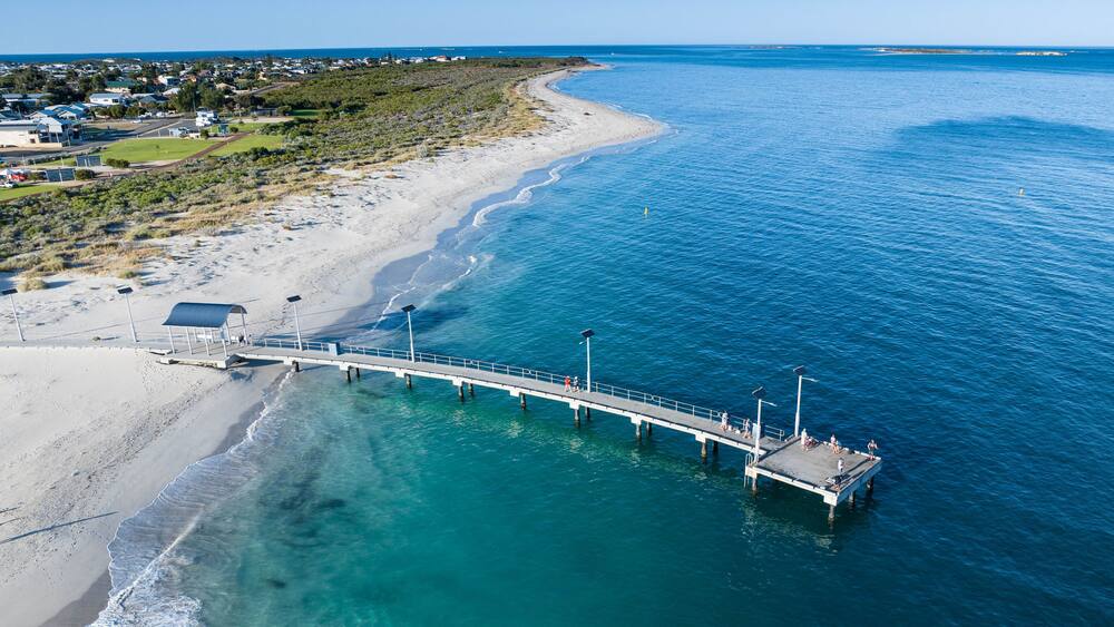 Une jetée s’étendant dans des eaux turquoise, une plage de sable et un quartier résidentiel en arrière-plan.