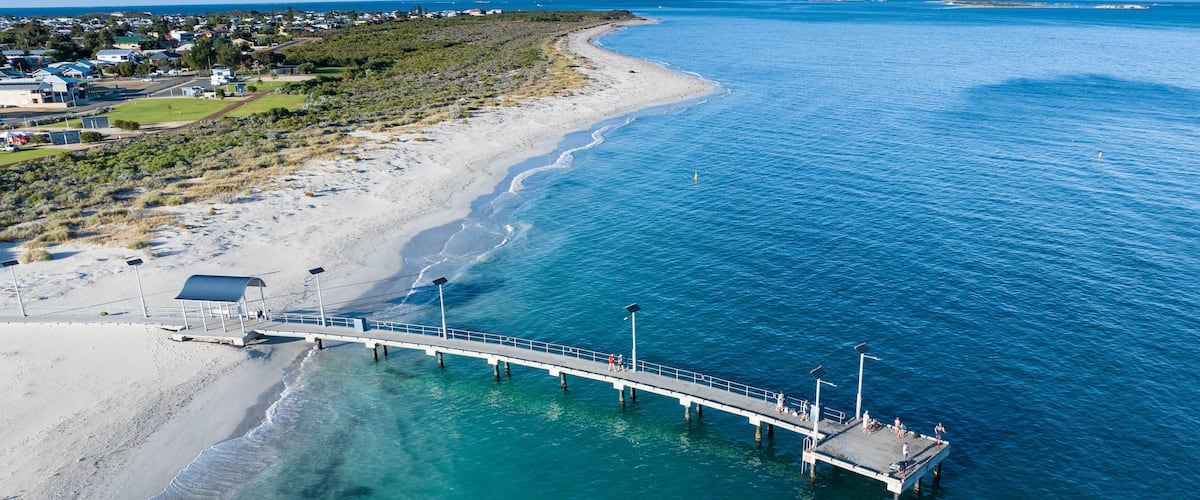 A coastal pier extending into turquoise waters, a sandy beach, and a residential area in the background.