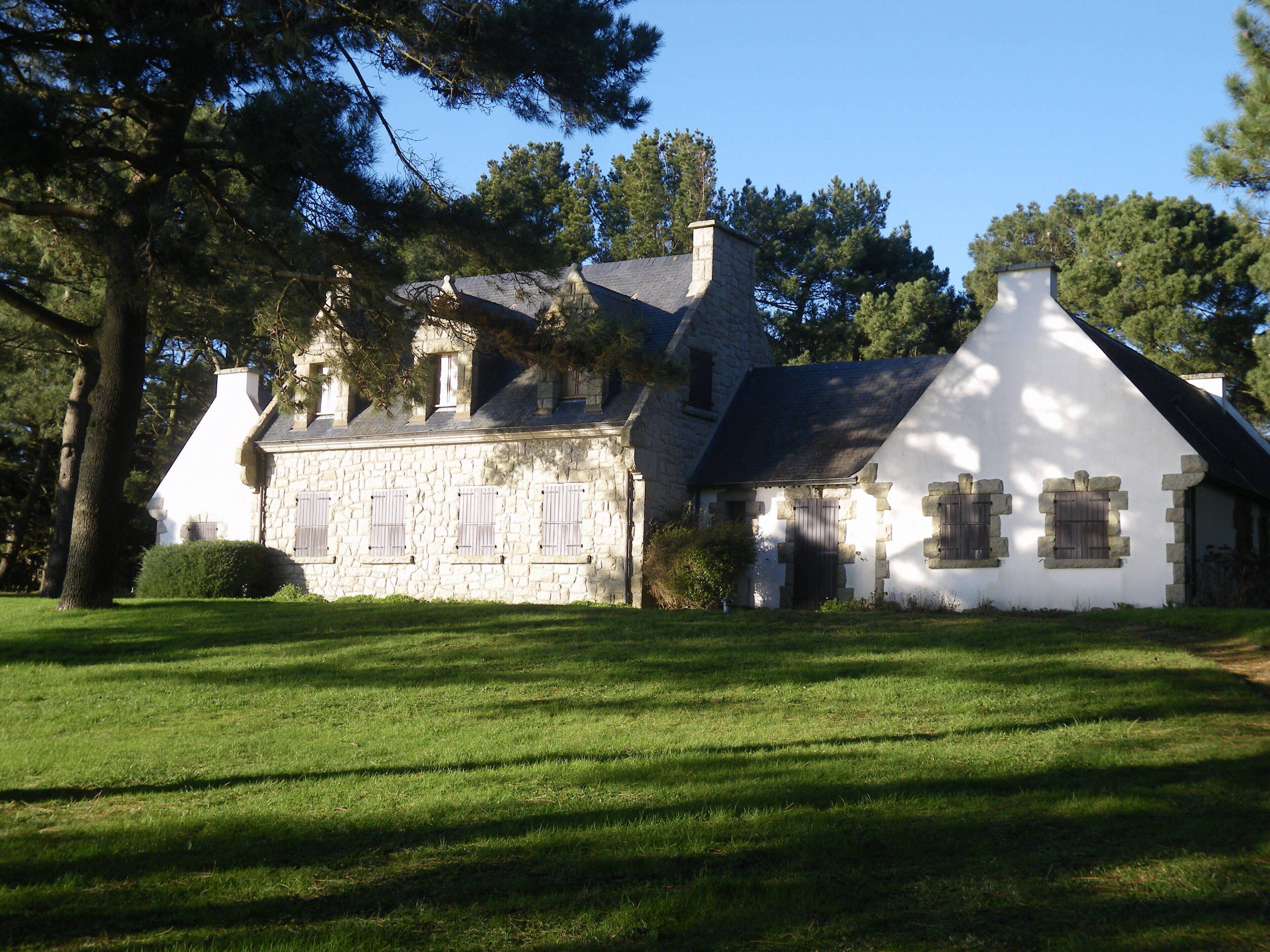 A stone house with a white facade and multiple chimneys, surrounded by a well-maintained lawn and tall trees.