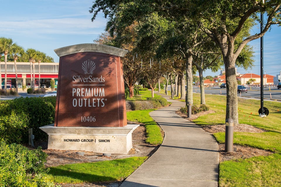 A SilverSands Premium Outlets sign with a pathway lined by trees and a road in the background.