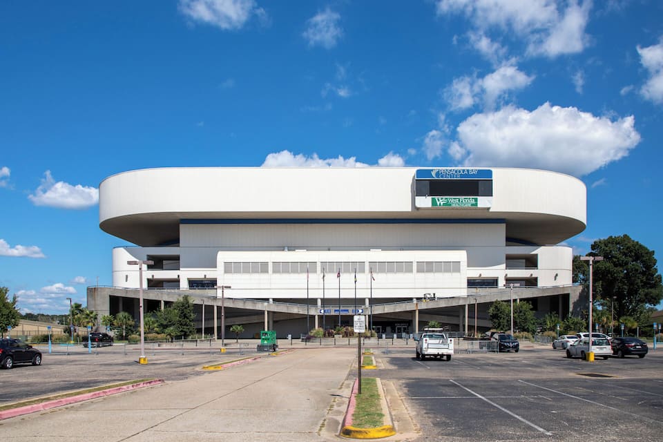Un edificio moderno con techo curvo y varios niveles, rodeado por un estacionamiento y un cielo azul despejado.