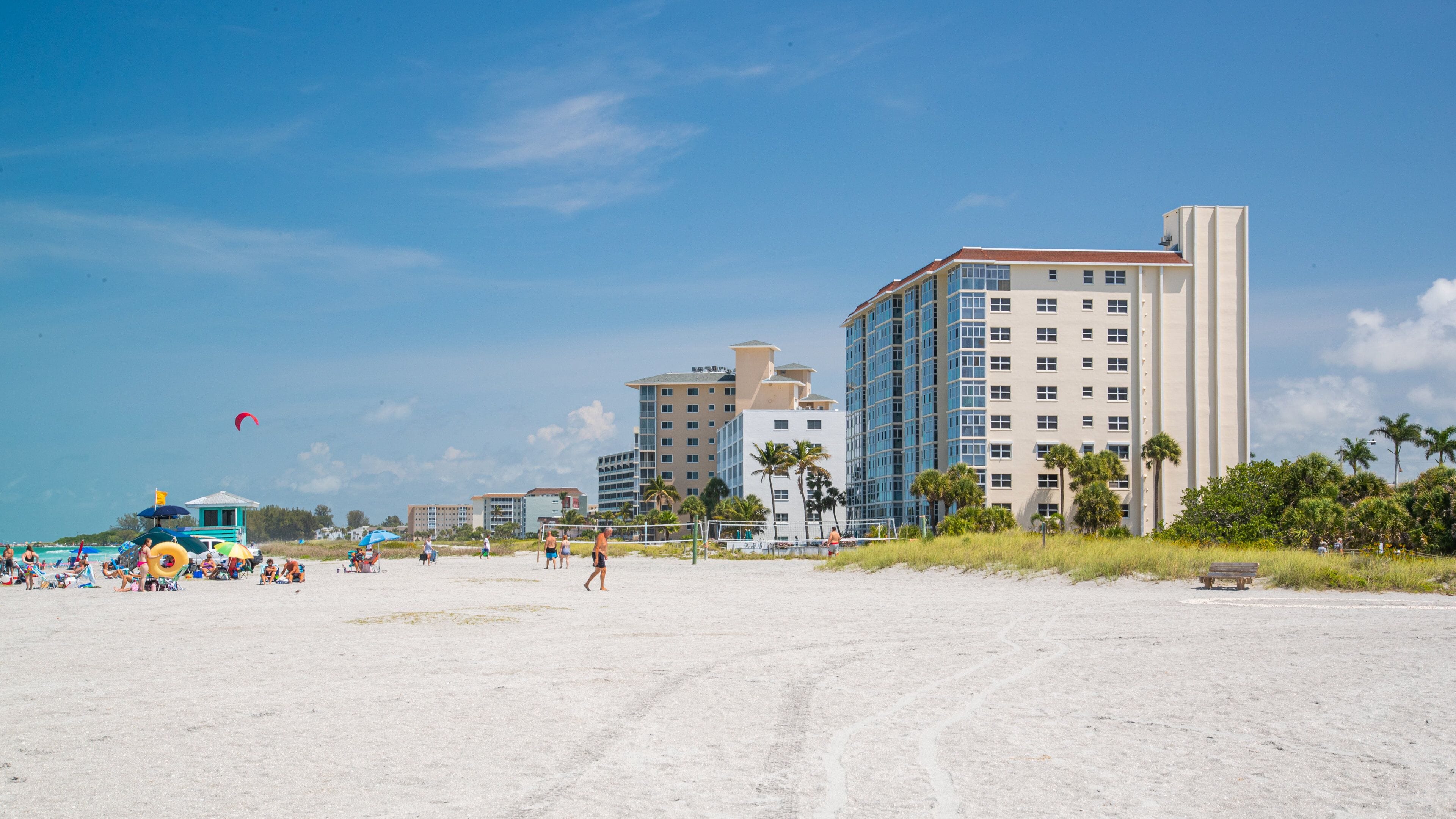 A sandy beach with people, a volleyball net, and tall buildings in the background.