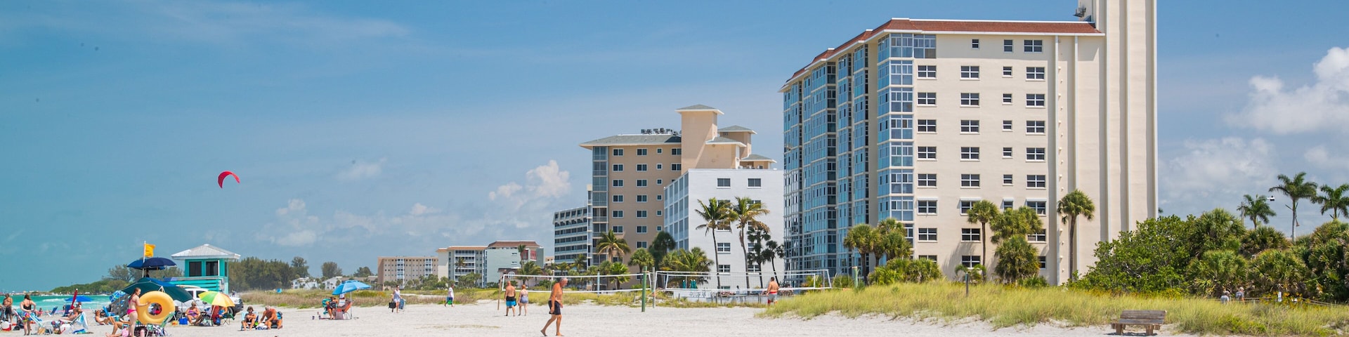 Uma praia de areia com pessoas, uma rede de voleibol e edifícios altos ao fundo.