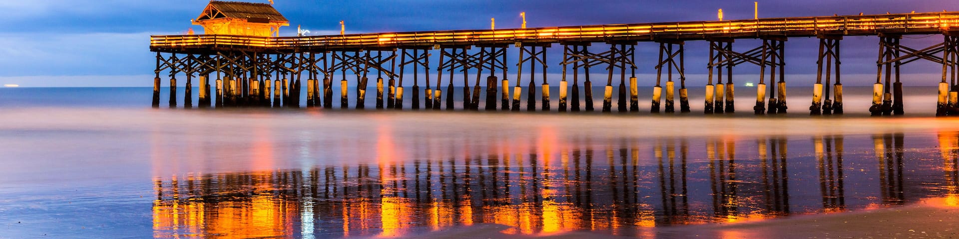 A pier extending into the water with a building at the end, surrounded by a calm sea and a cloudy sky.