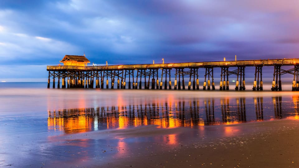 A pier extending into the water with a building at the end, surrounded by a calm sea and a cloudy sky.