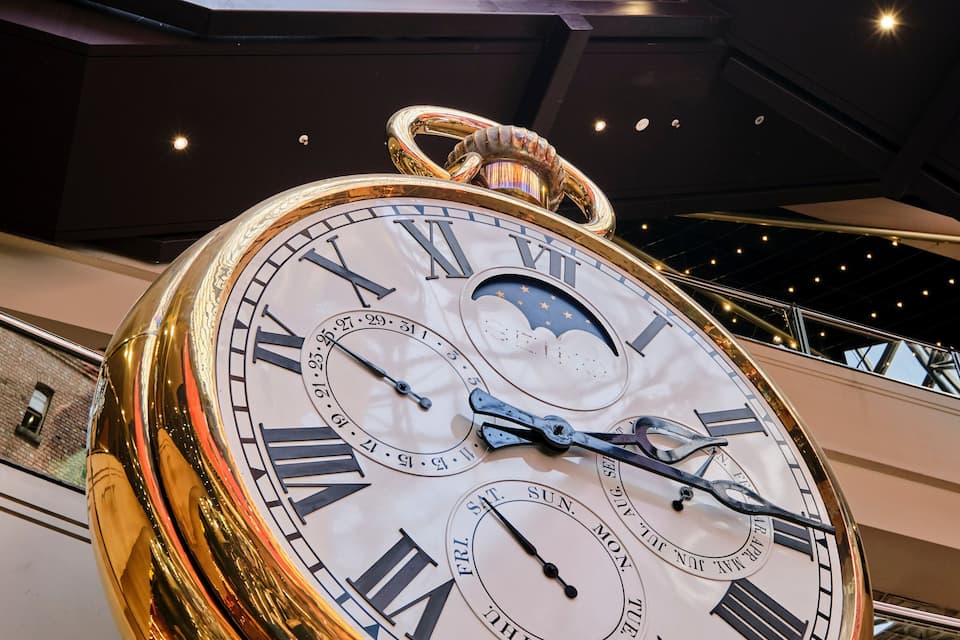 A large, ornate clock with Roman numerals and multiple dials, suspended in a modern building with a glass ceiling.