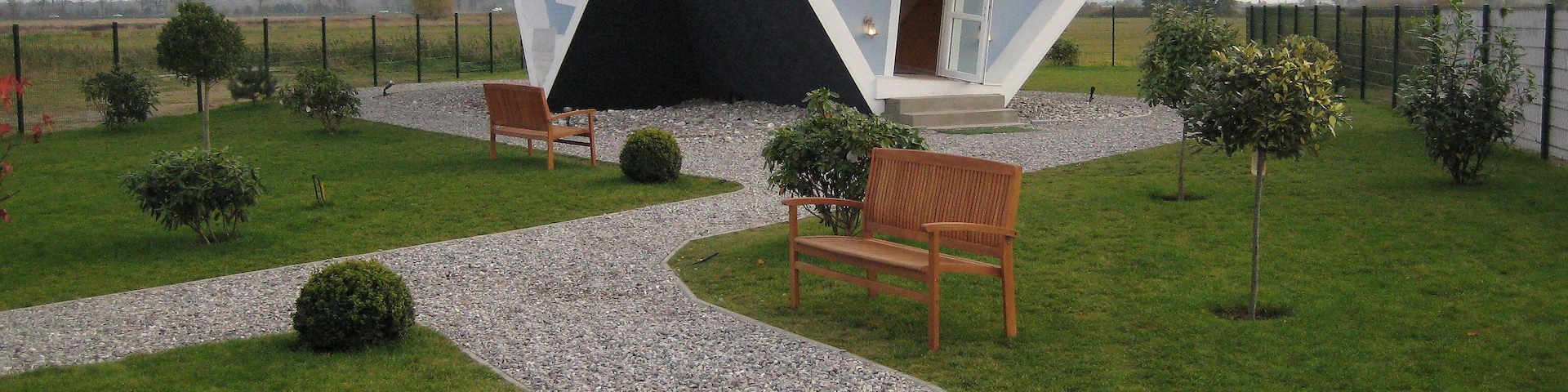 Una casa moderna con un diseño arquitectónico único, rodeada de un jardín cuidado con senderos, bancos y pequeños árboles.