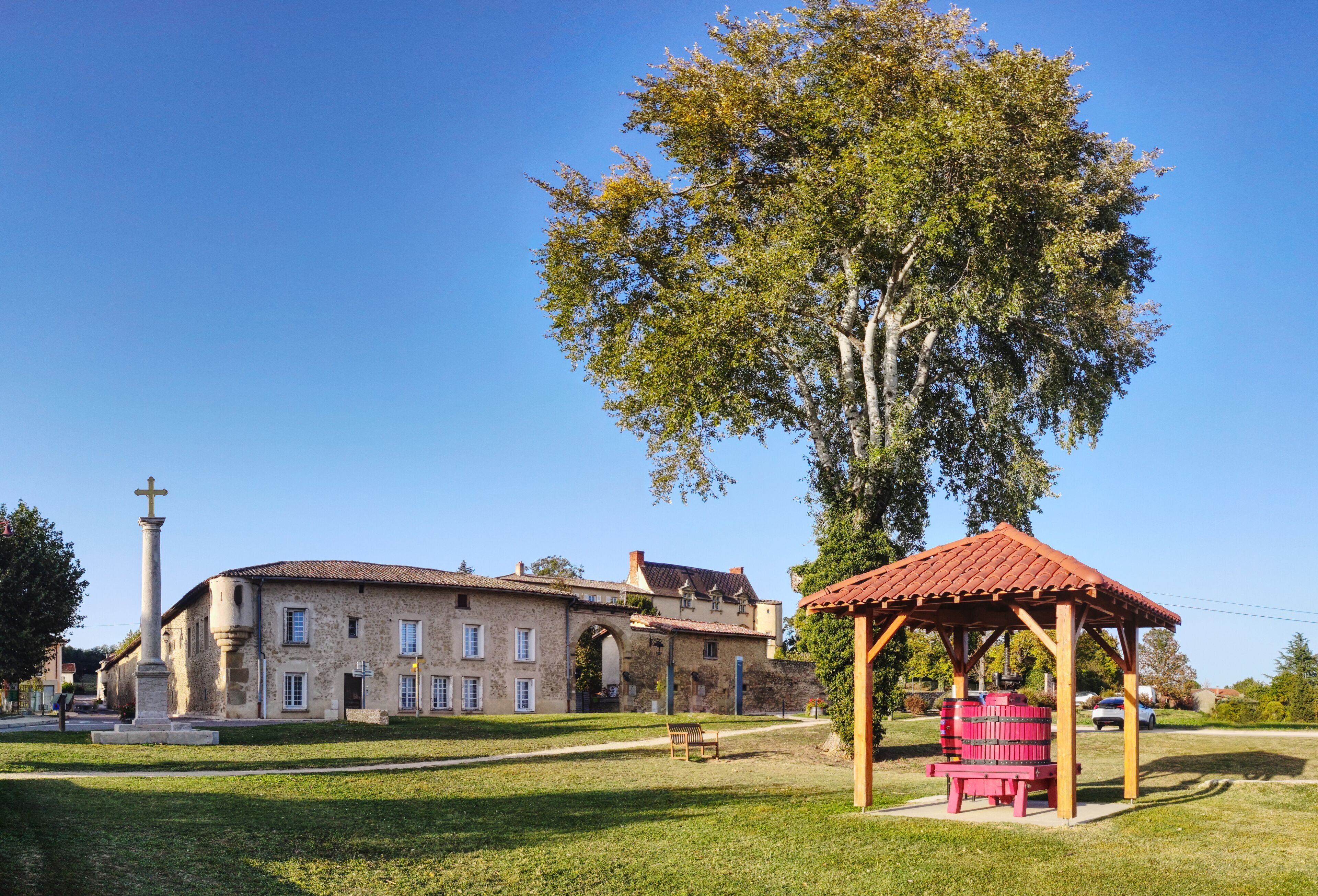 A historic stone building with a large tree in front, a wooden gazebo, and a cross monument.