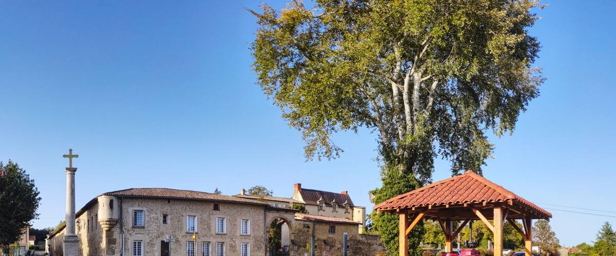 A historic stone building with a large tree in front, a wooden gazebo, and a cross monument.