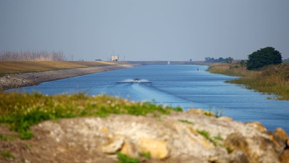 A river with a wake behind a boat, surrounded by grassy banks and a distant industrial structure.
