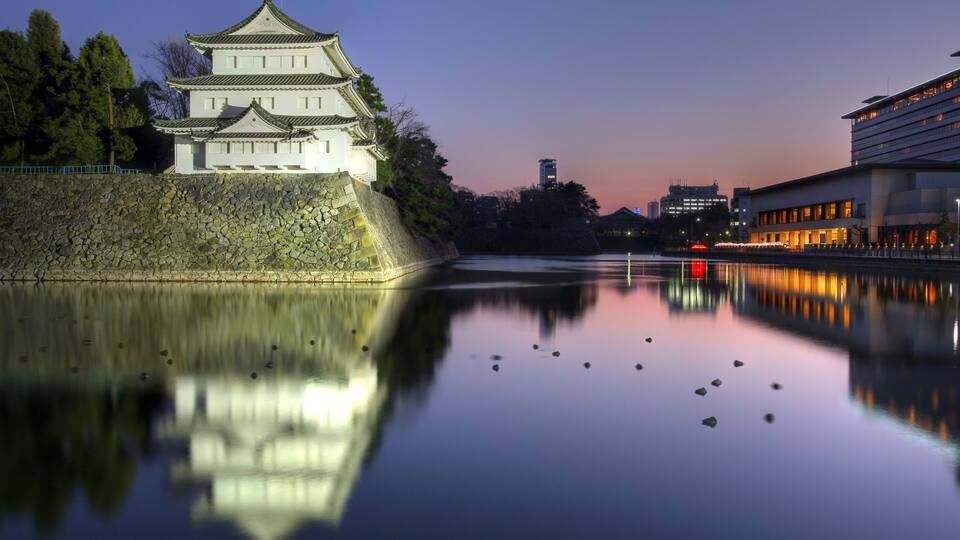 A traditional Japanese castle by a calm river with modern buildings in the background.