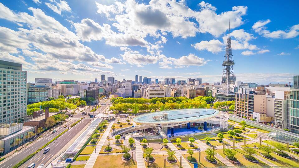 A cityscape with modern buildings, a prominent tower, and a large circular plaza with trees.