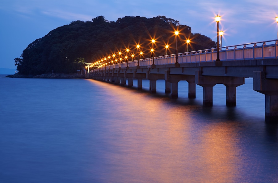 A long bridge with streetlights extending over calm water, leading to a forested island.