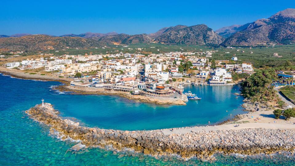 A coastal town with clear blue waters, a rocky breakwater, and mountains in the background.