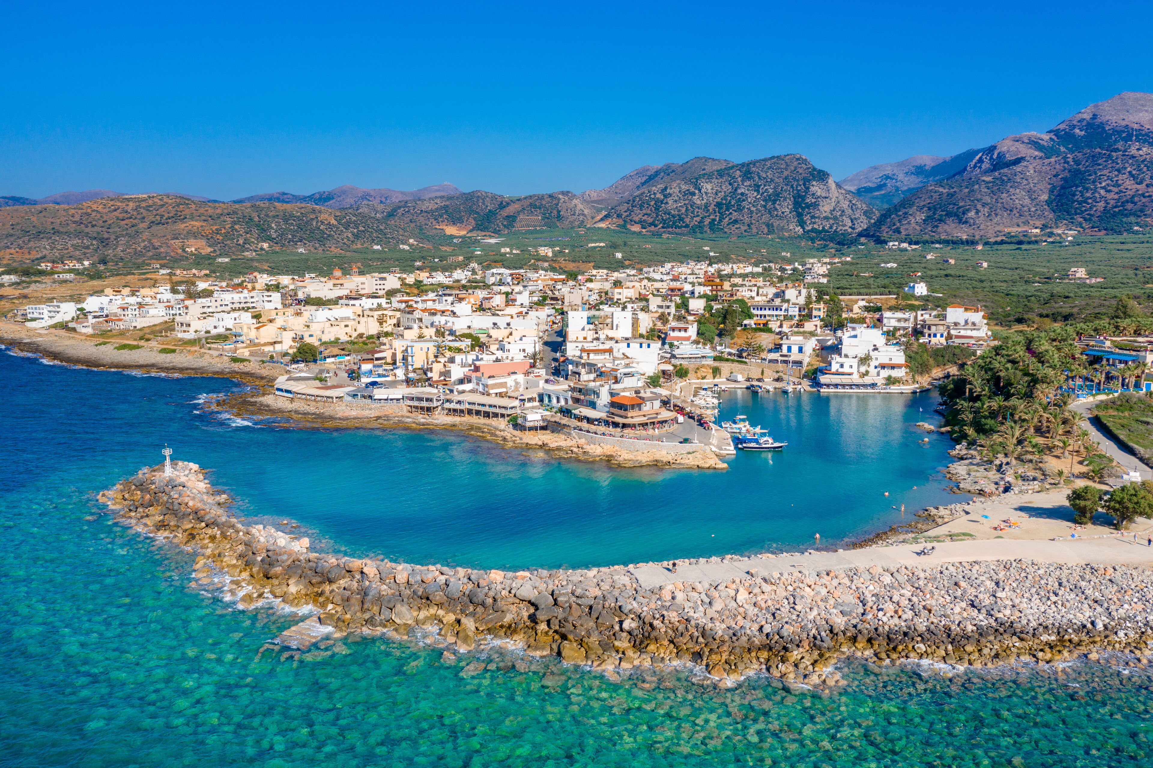 A coastal town with clear blue waters, a rocky breakwater, and mountains in the background.