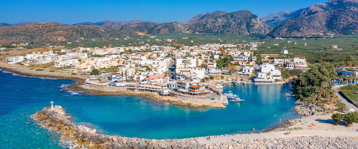 A coastal town with clear blue waters, a rocky breakwater, and mountains in the background.