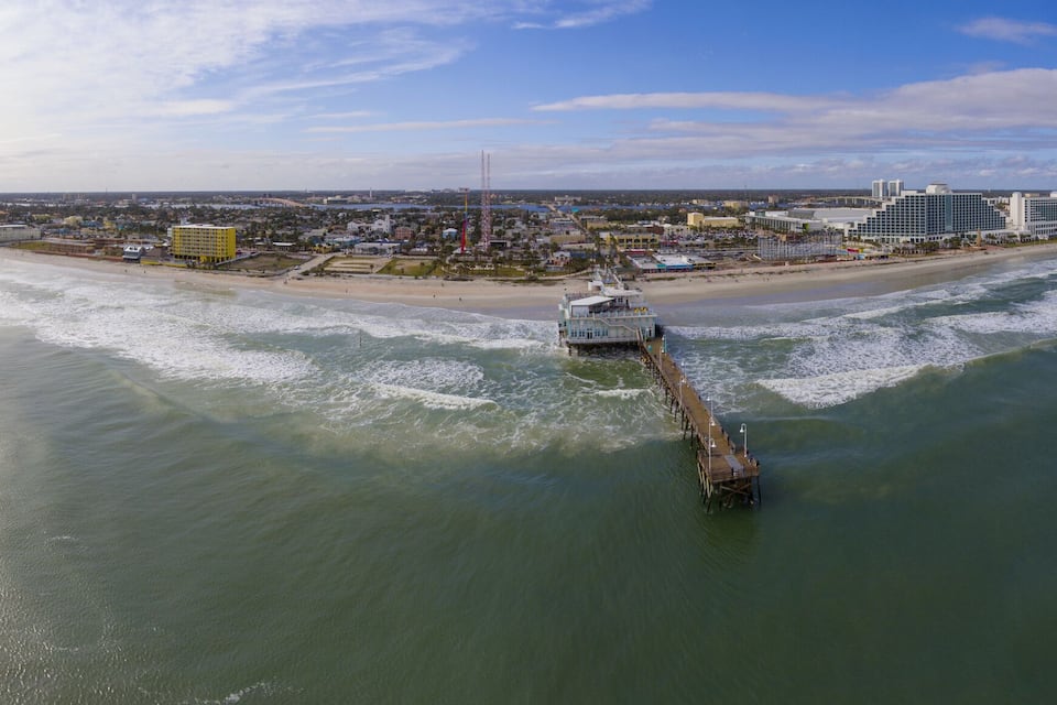An aerial view of a coastal city with a pier extending into the ocean, buildings along the shore, and a clear sky.