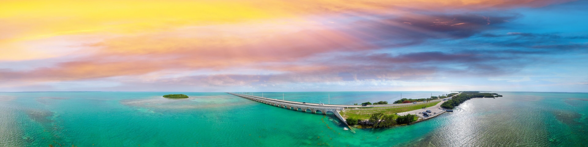 Uma ponte liga duas massas de terra sobre águas turquesa, rodeada por céus límpidos e um pôr do sol.