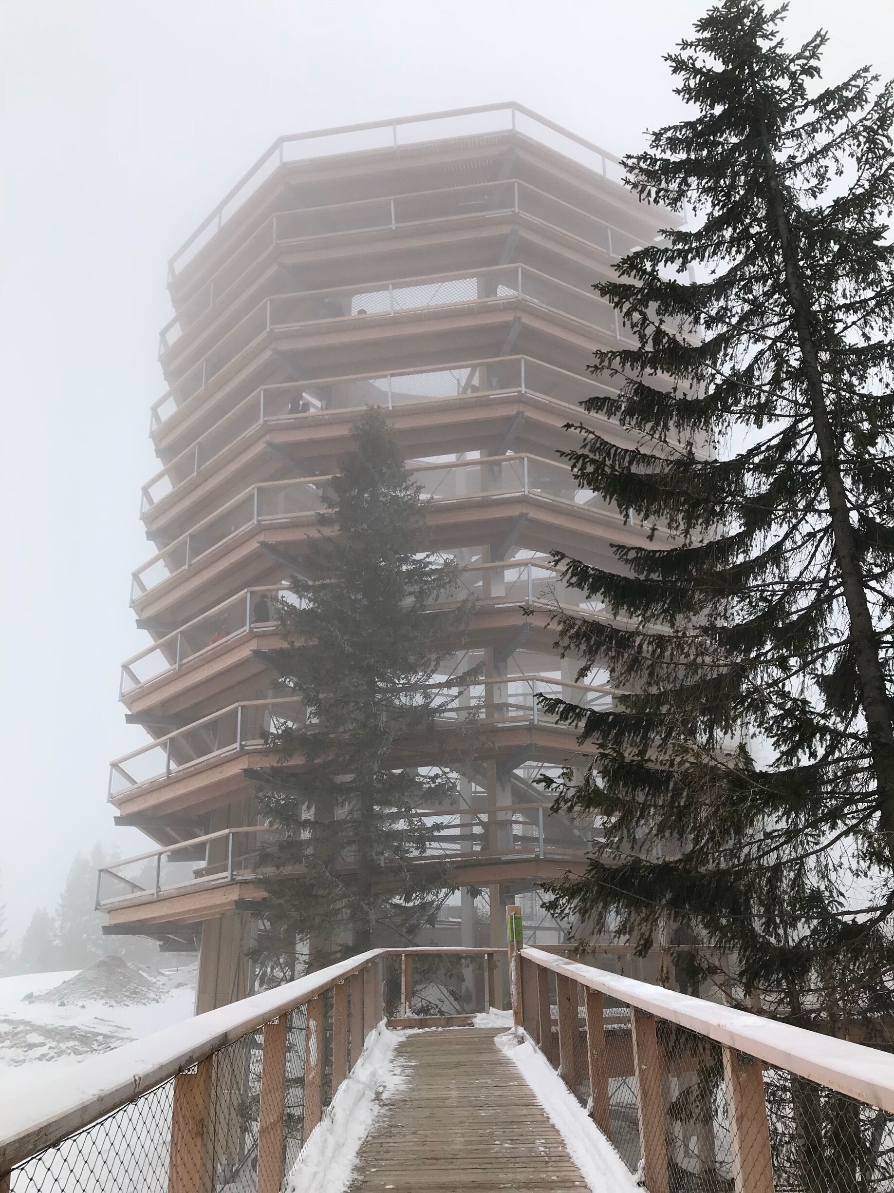 A multi-story building with a wooden deck leading up to it, surrounded by snow and trees.