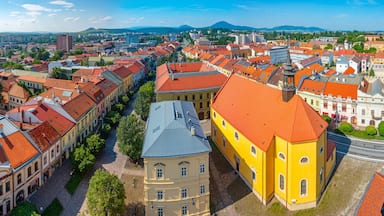 Paisagem urbana com edifícios de telhado vermelho, um edifício amarelo em destaque e uma cadeia montanhosa ao longe.