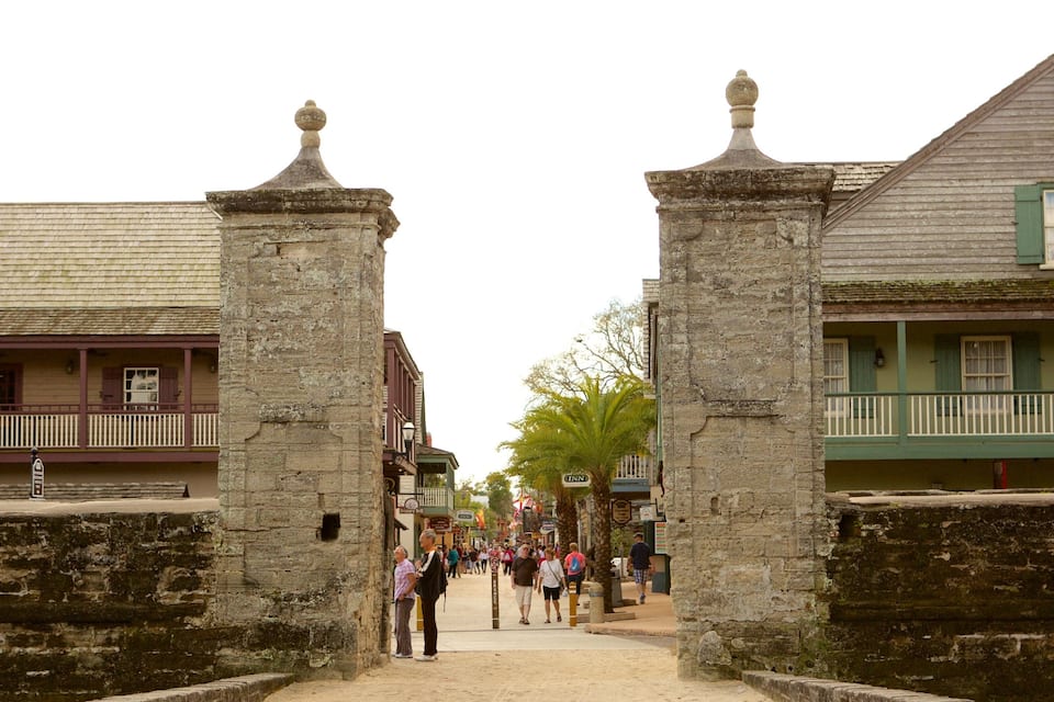 A historic street with stone gateposts, colonial buildings, and pedestrians.