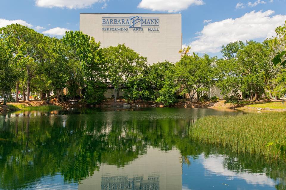 A serene pond with a reflection of trees and a building labeled "Barbara Mann Performing Arts Hall".