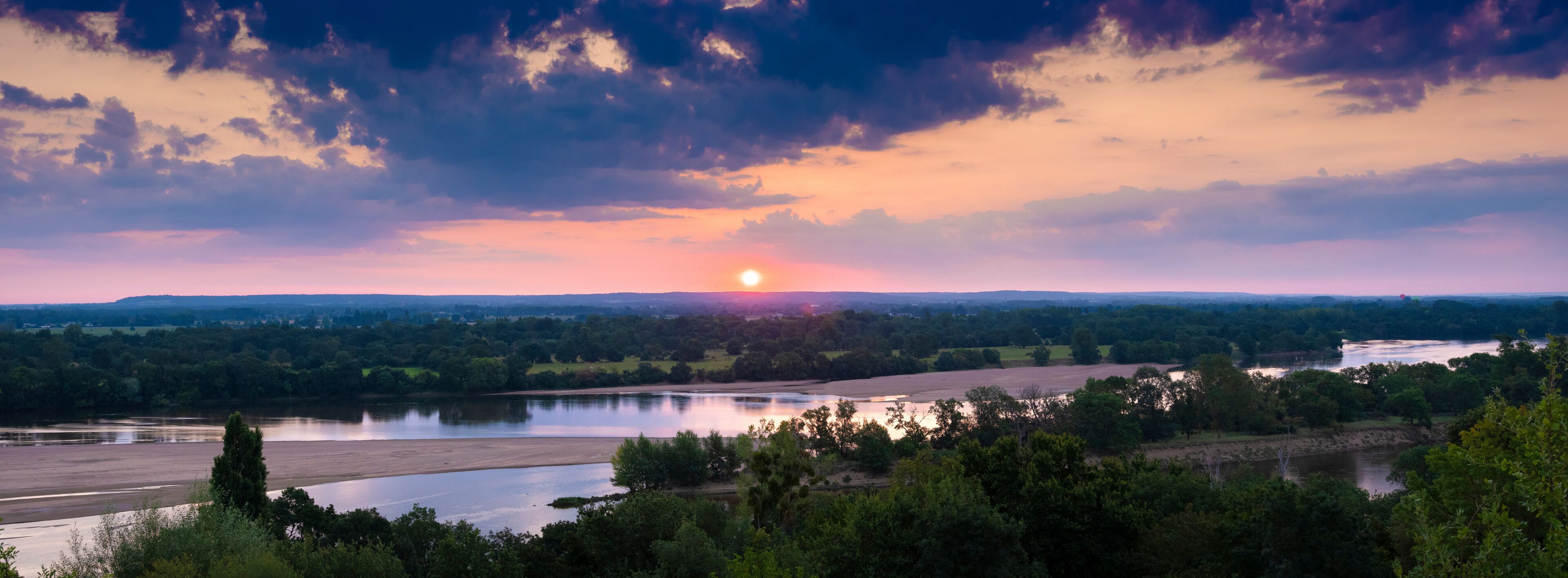 Pôr do sol sobre um vale fluvial com paisagem arborizada e colinas ao longe.