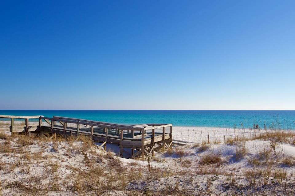 Una pasarela de madera que conduce a una playa con aguas azules cristalinas y dunas de arena.