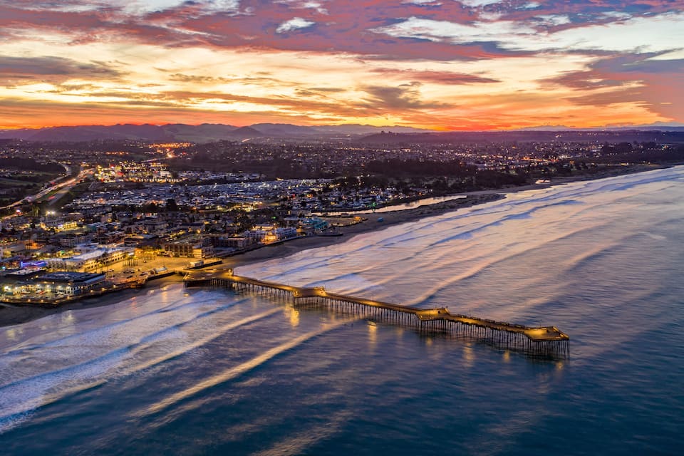 Uma cidade costeira ao pôr do sol, com um cais que se estende para o oceano, um calçadão iluminado e montanhas ao fundo.