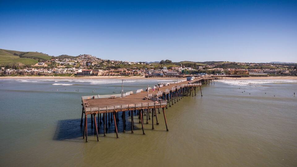 Un muelle se adentra en una playa tranquila con un paisaje de un pueblo costero y colinas.