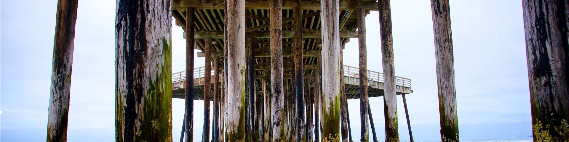 A weathered pier with wooden beams and columns, extending over a sandy beach with calm water.